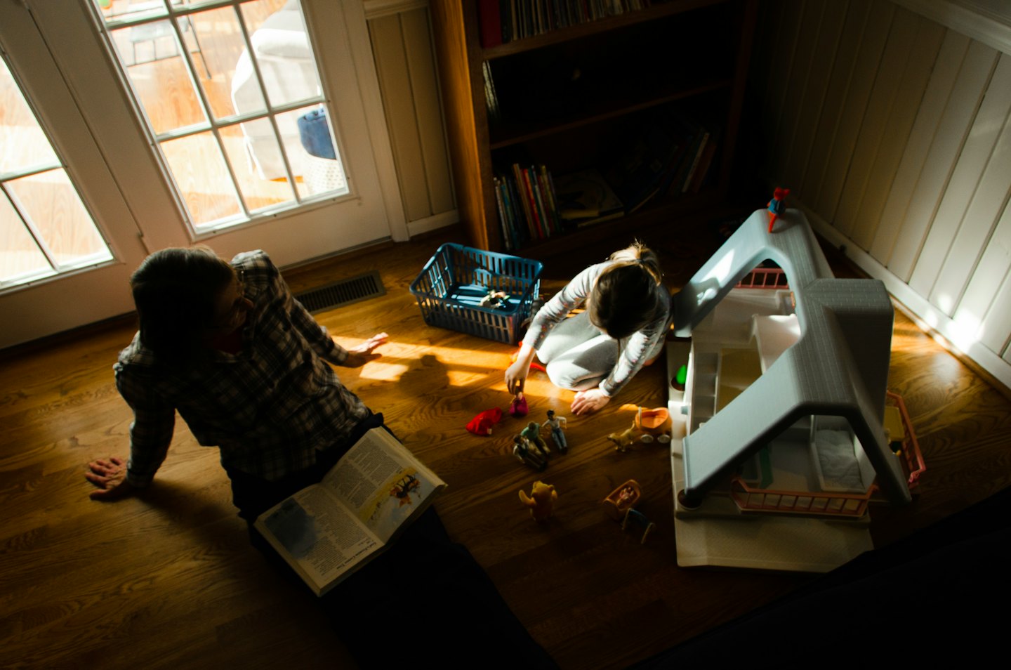 Adult reads a book while child plays with dollhouse on floor.
