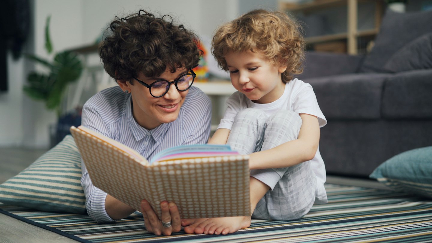 Woman and boy read a colorful picture book on a rug.